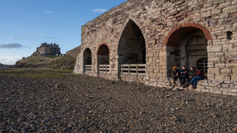 Group of people sitting inside the Limekilns, Lindisfarne Castle in the background.